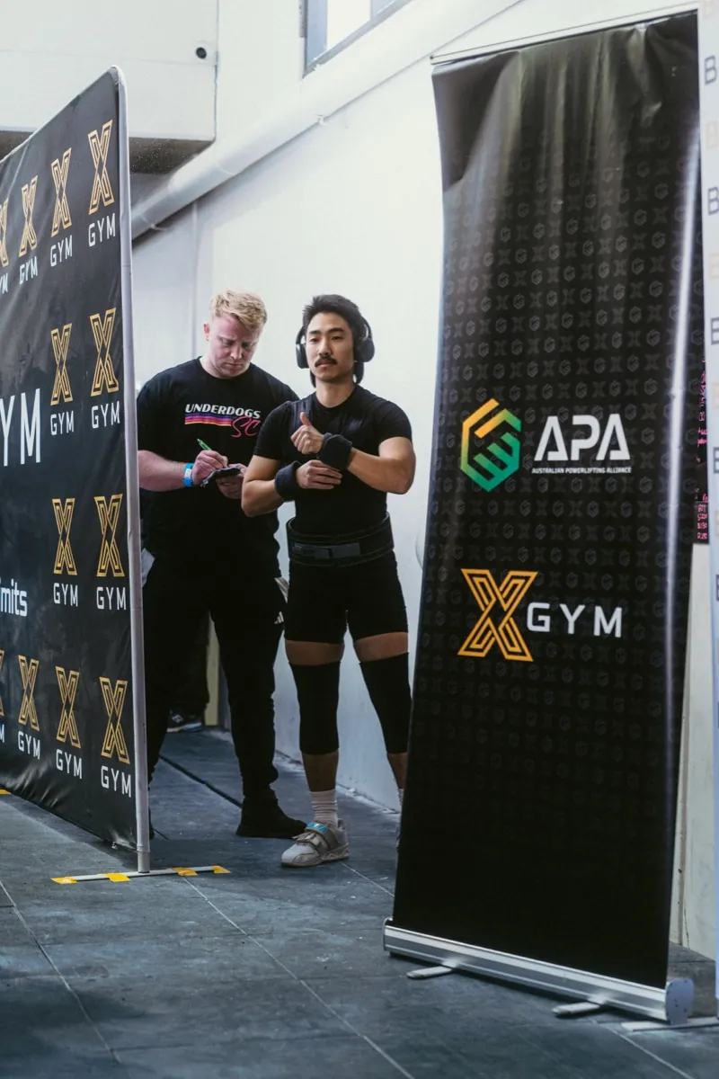 A coach writing down attempts beside a lifter warming up backstage