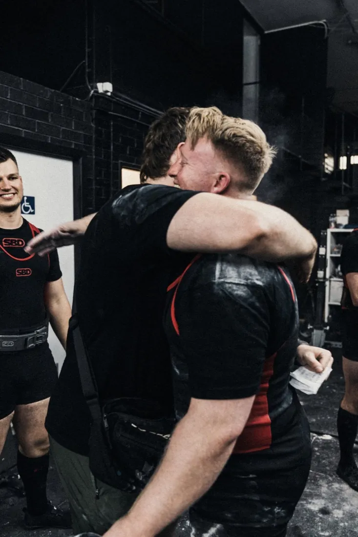 Two lifters hugging in a chalk-filled warm-up room after a successful attempt